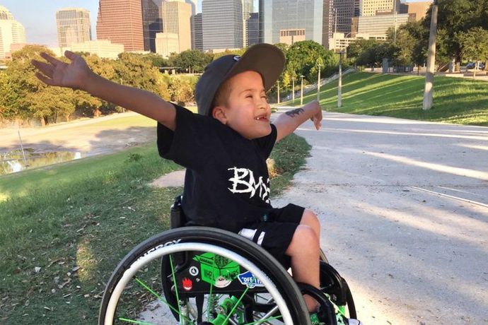 Boy in wheelchair with arms outstretched, smiling. He's in a park, downtown buildings in the background.
