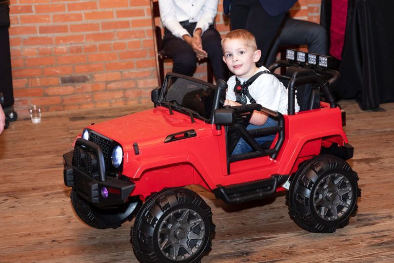 Young boy smiles while driving a red toy Jeep indoors.
