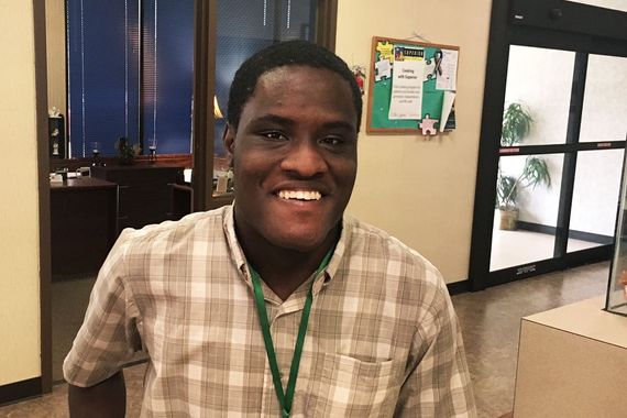 Smiling  man wearing a plaid shirt and lanyard stands indoors near a bulletin board and glass doors.