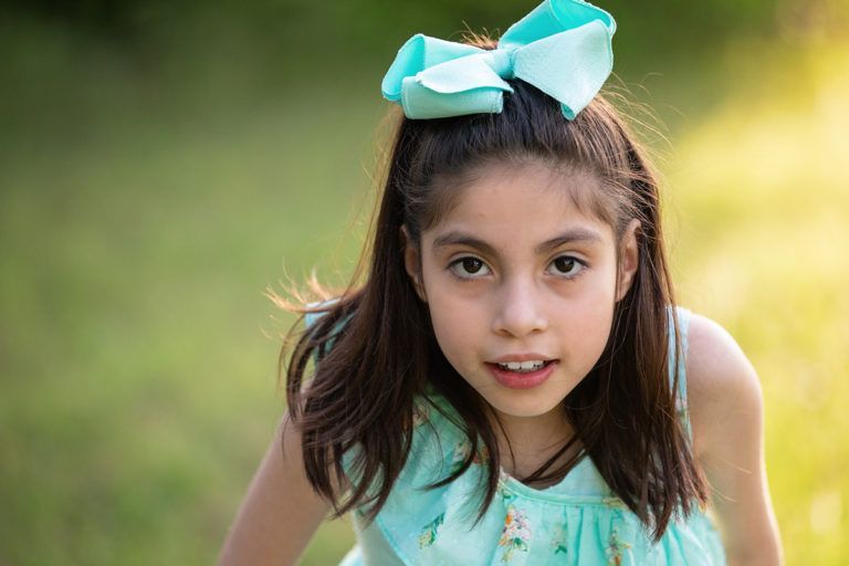 Girl with brown hair and turquoise bow smiles outdoors.