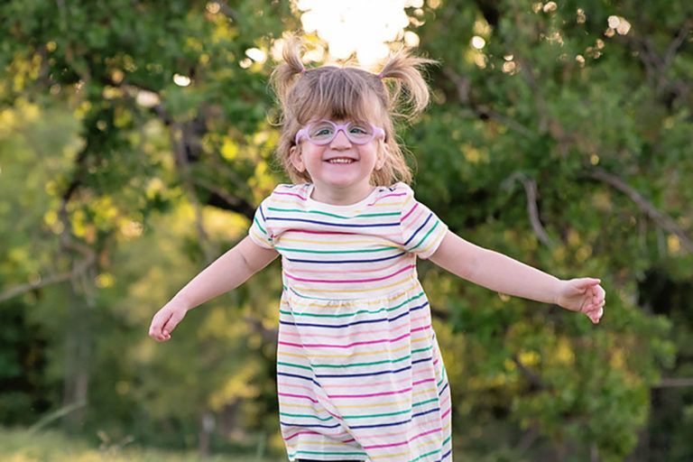 Young girl with pigtails, glasses, and a striped dress smiles broadly outdoors with arms outstretched.