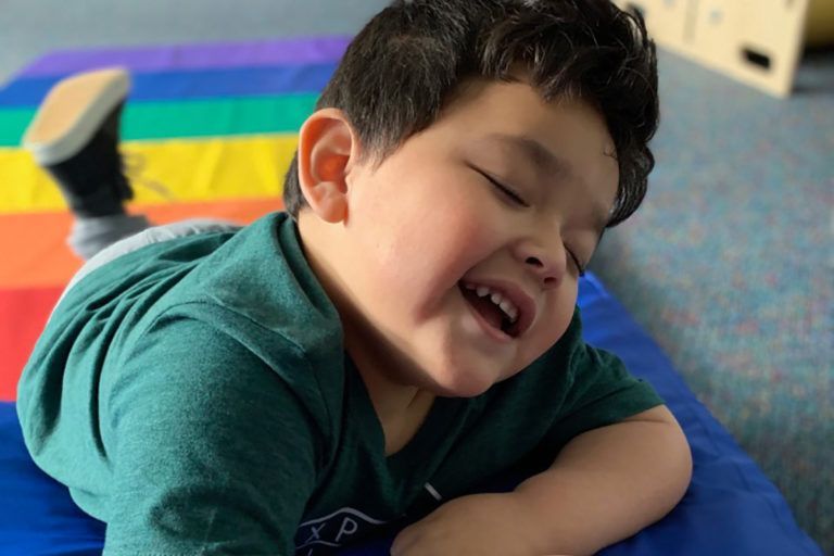 Smiling child lying on a blue mat, with a rainbow mat in the background.