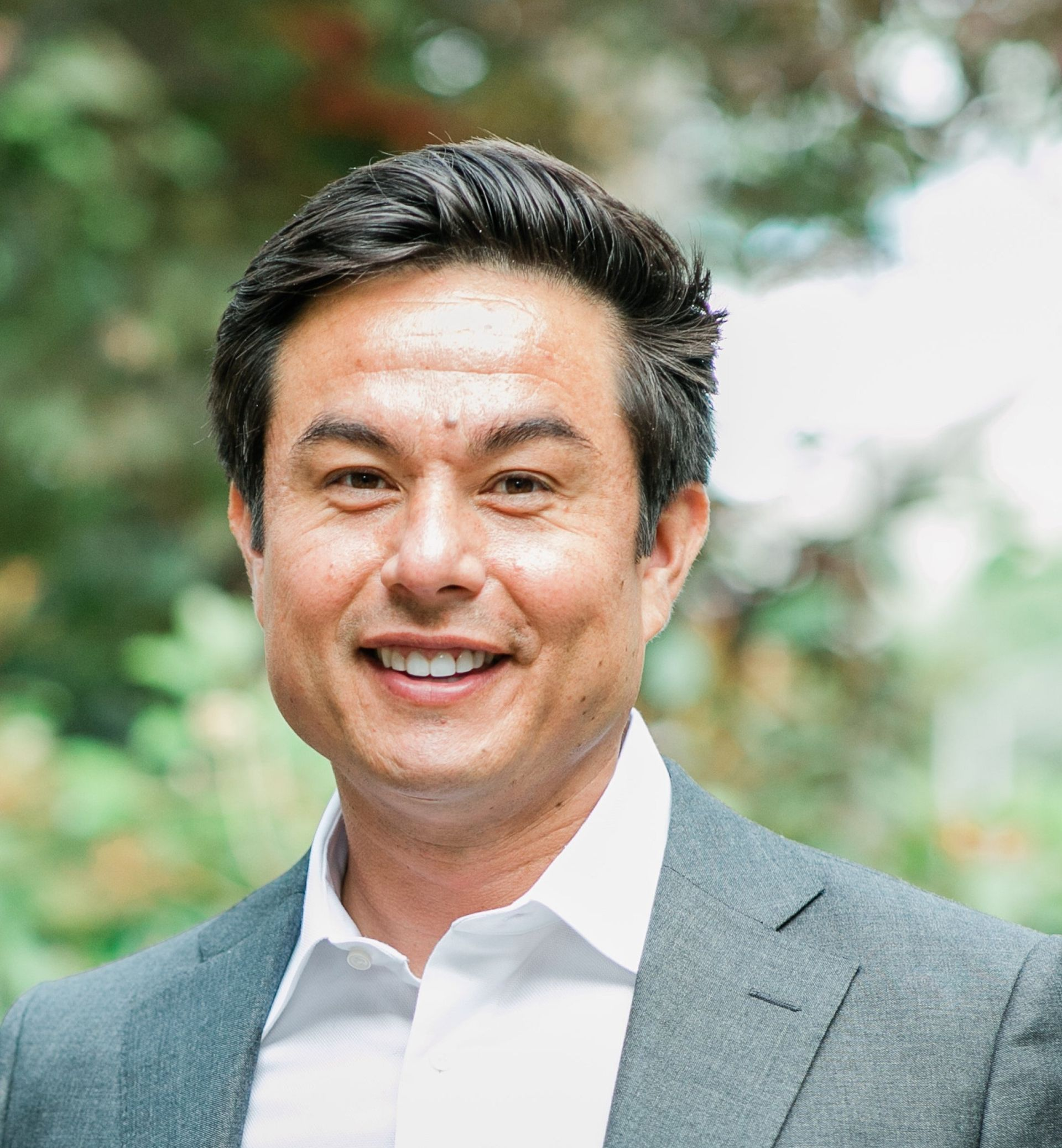 Smiling man in a gray blazer and white shirt outdoors with blurred greenery behind him
