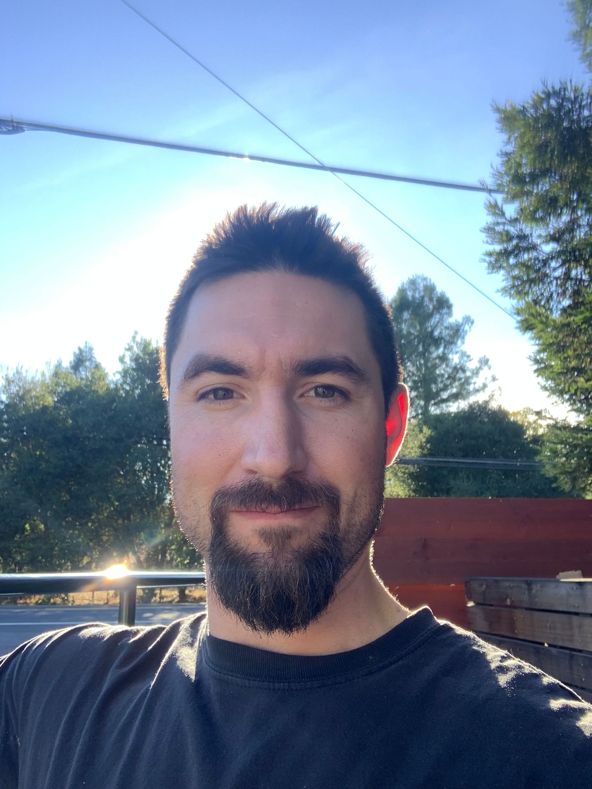 Man standing outdoors in sunlight with trees and a blue sky in the background