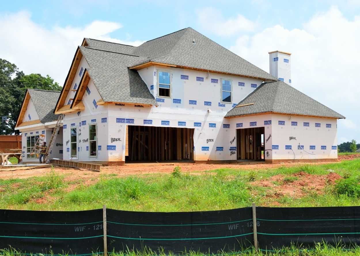 A new house under construction, with gray roof shingles and white weather-resistant exterior siding, sits on a dirt lot.