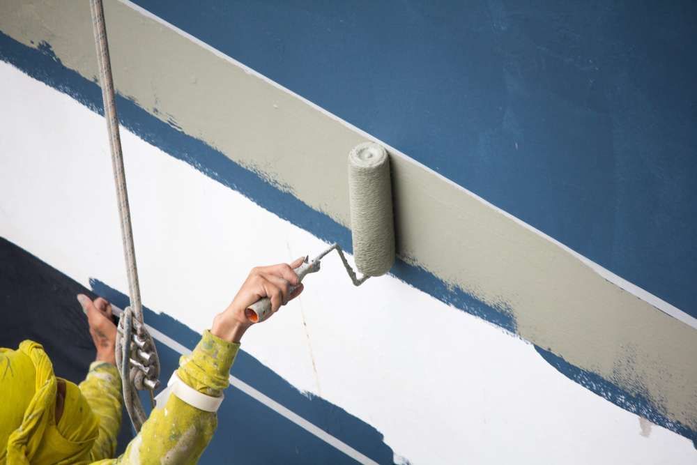 A painter in a yellow top uses a roller to apply beige paint to a blue wall, suspended by a rope.