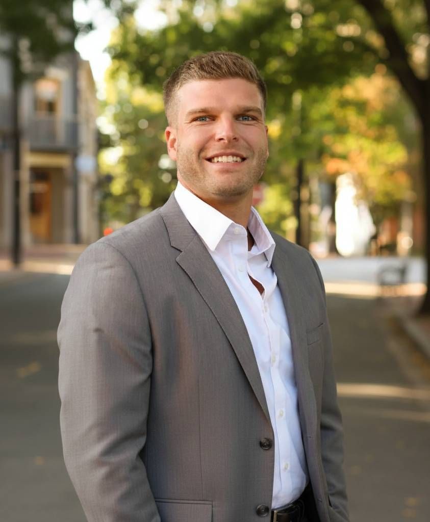 A smiling person wearing a grey suit jacket and white button-down shirt, standing outdoors in a park-like setting.