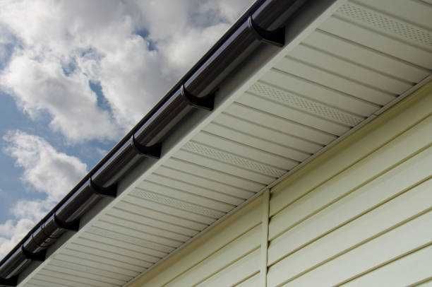 A low-angle view of a house exterior showing light yellow siding, white vented soffit, and a dark brown rain gutter.