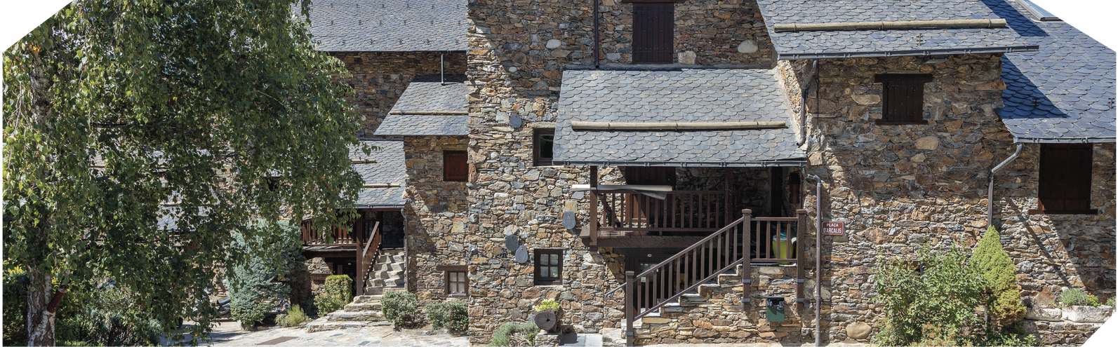 Edifici de pedra amb diversos nivells, teulades grises i detalls de fusta, situat enmig de vegetació. Premis PAA.
