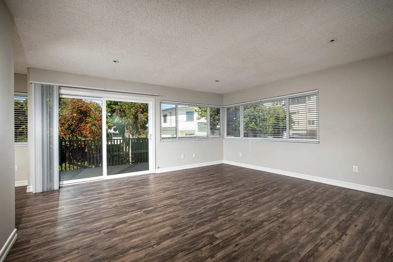 Living room with large sliding glass doors and multiple windows. Dark wood-look flooring and light grey walls.