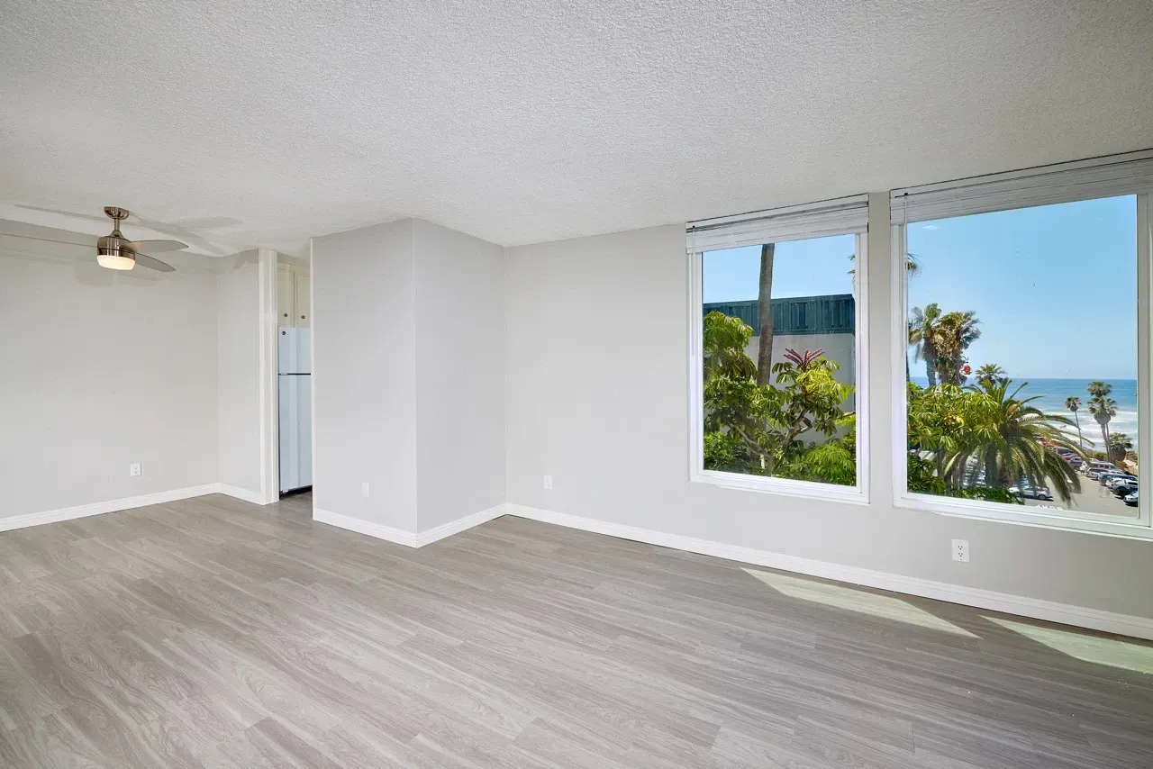 Empty living room with wood floors, ceiling fan, and large windows overlooking the ocean and palm trees.