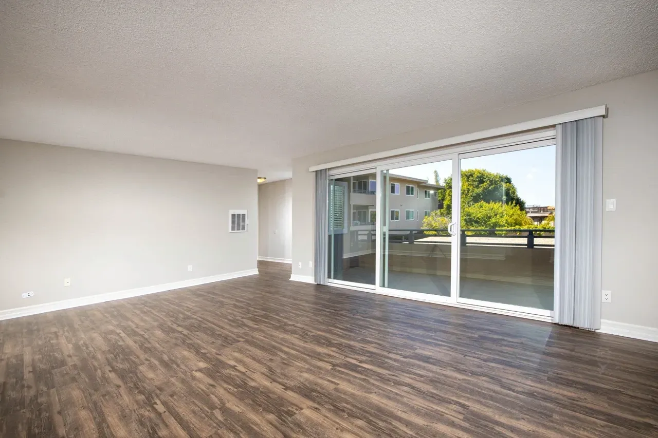 Empty living room with wood-look flooring and sliding glass doors overlooking a balcony and building exterior.