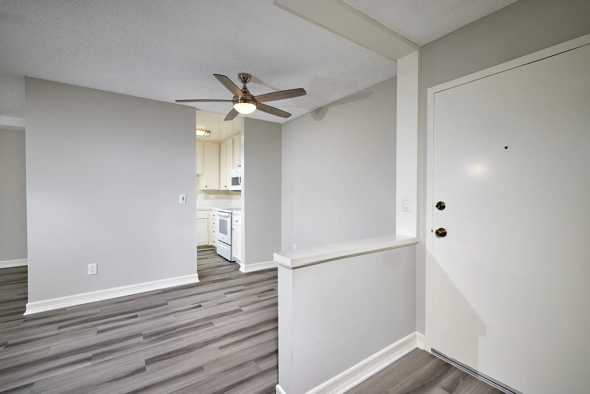 View of an apartment entryway with a white door, ceiling fan, and a glimpse of the kitchen.