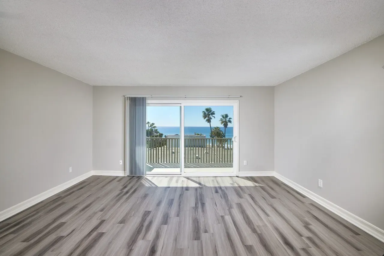 Empty living room with sliding glass doors opening to a balcony with ocean and palm tree views.