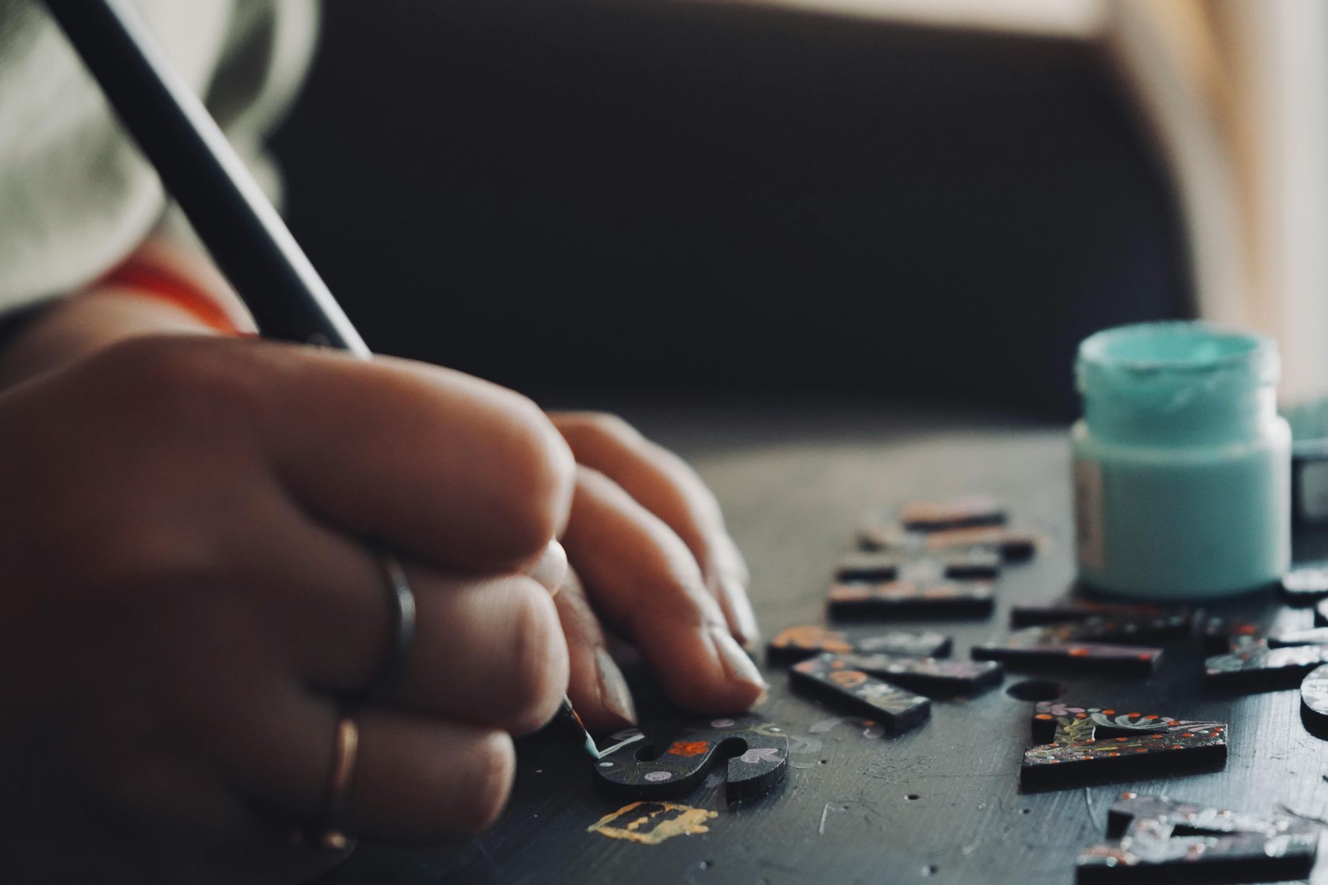 Hand painting small black pieces on a tabletop with a teal paint jar nearby