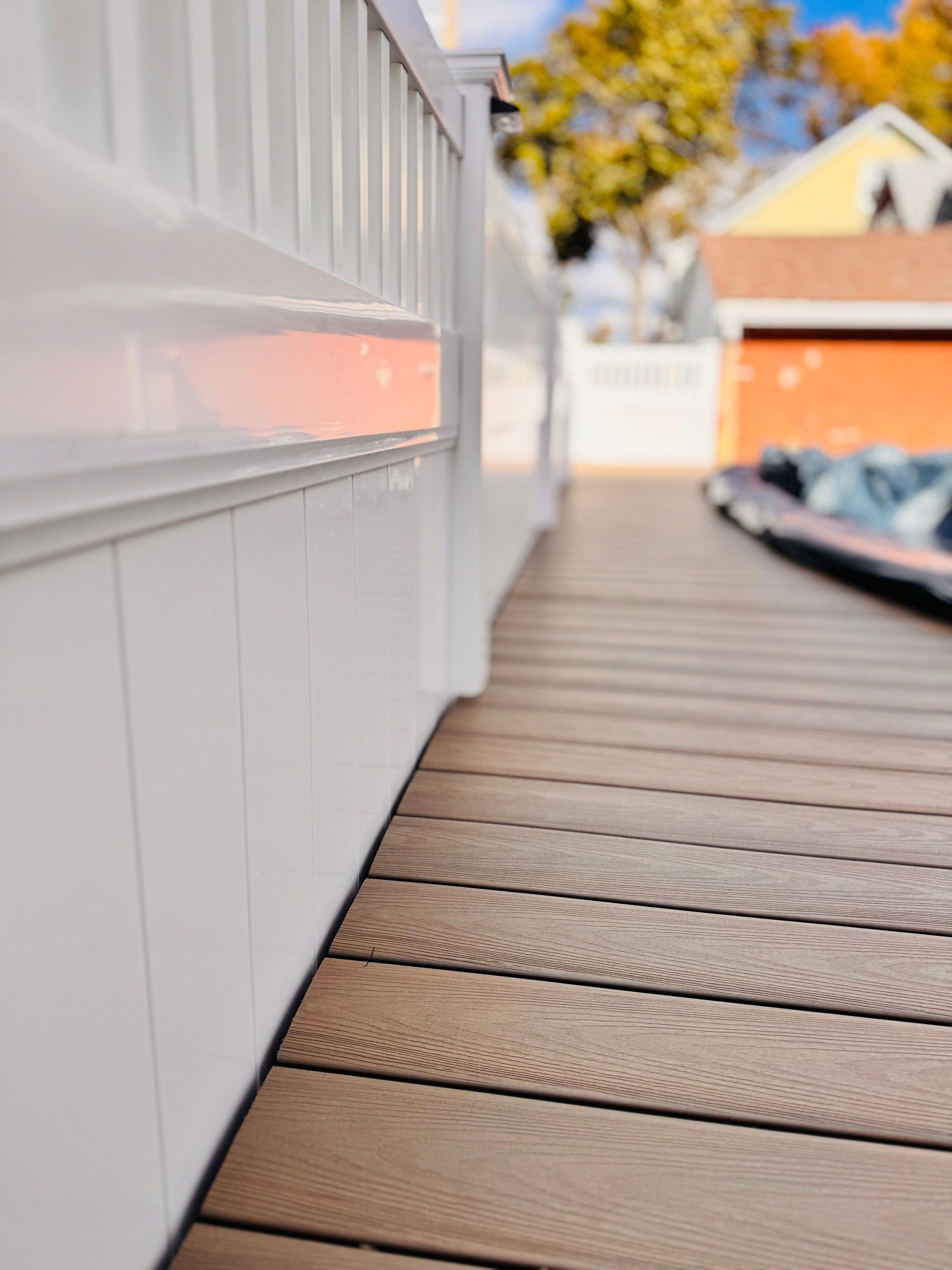 White deck railing and paneling bordering a composite wood deck with a blurred background.