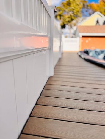 White deck railing and paneling bordering a composite wood deck with a blurred background.