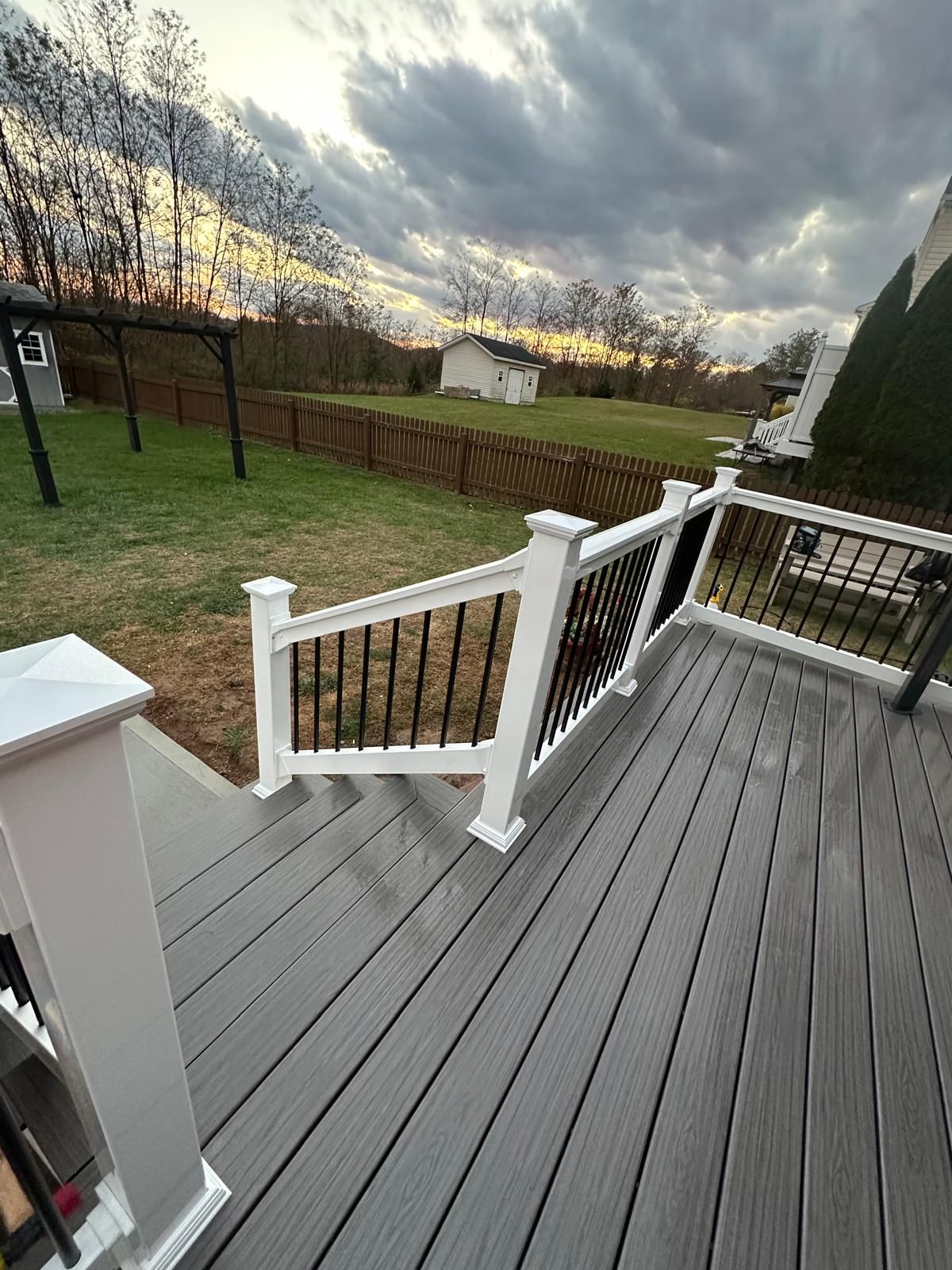 Deck with gray boards, white railing posts, and black spindles, overlooking a grassy yard under a cloudy sky.