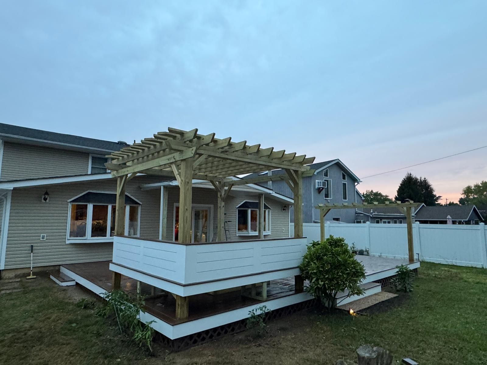 Wooden deck with pergola attached to a light-colored house at dusk.