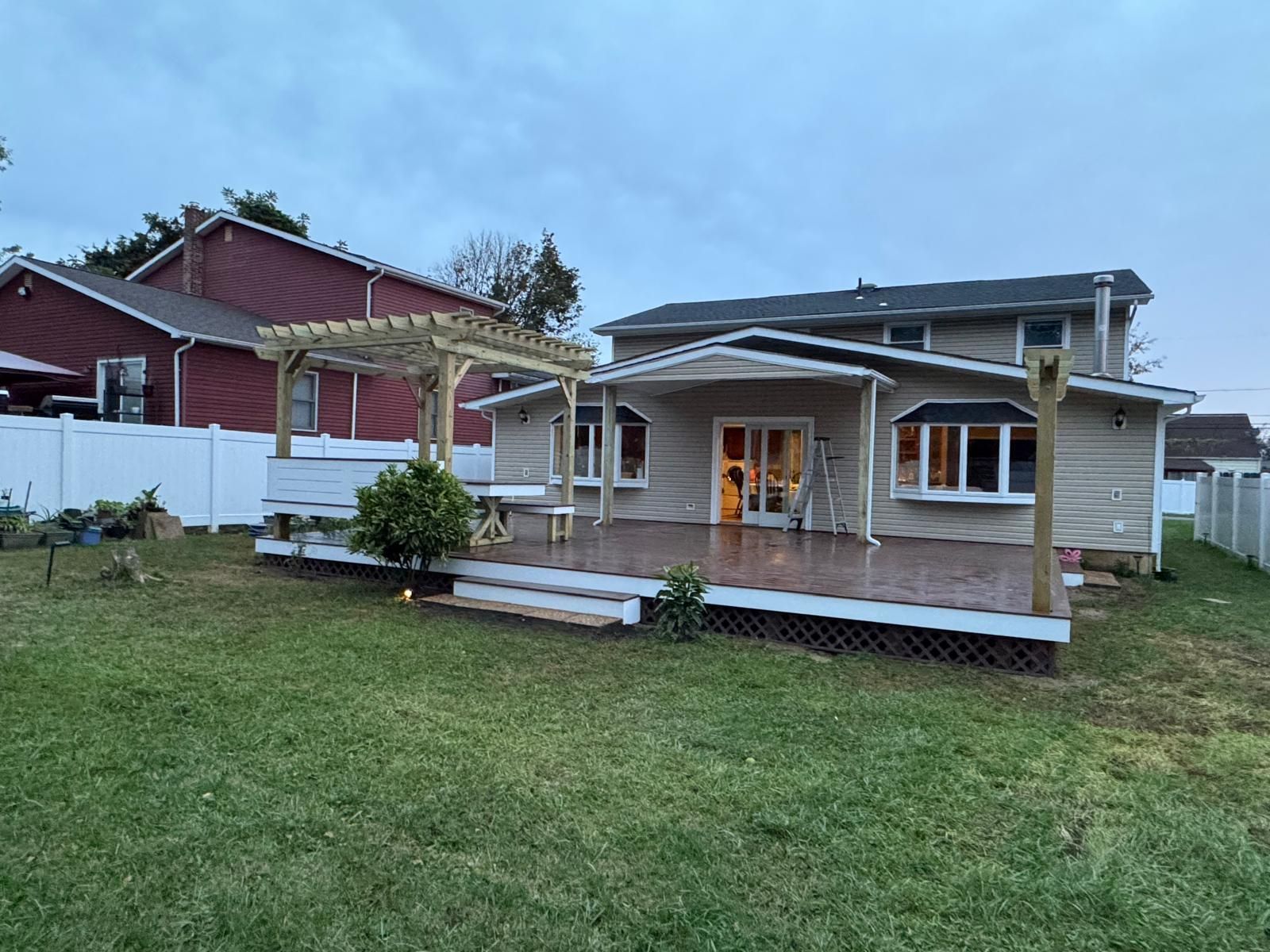 Backyard deck with pergola attached to a beige house, lawn in front, white fence.