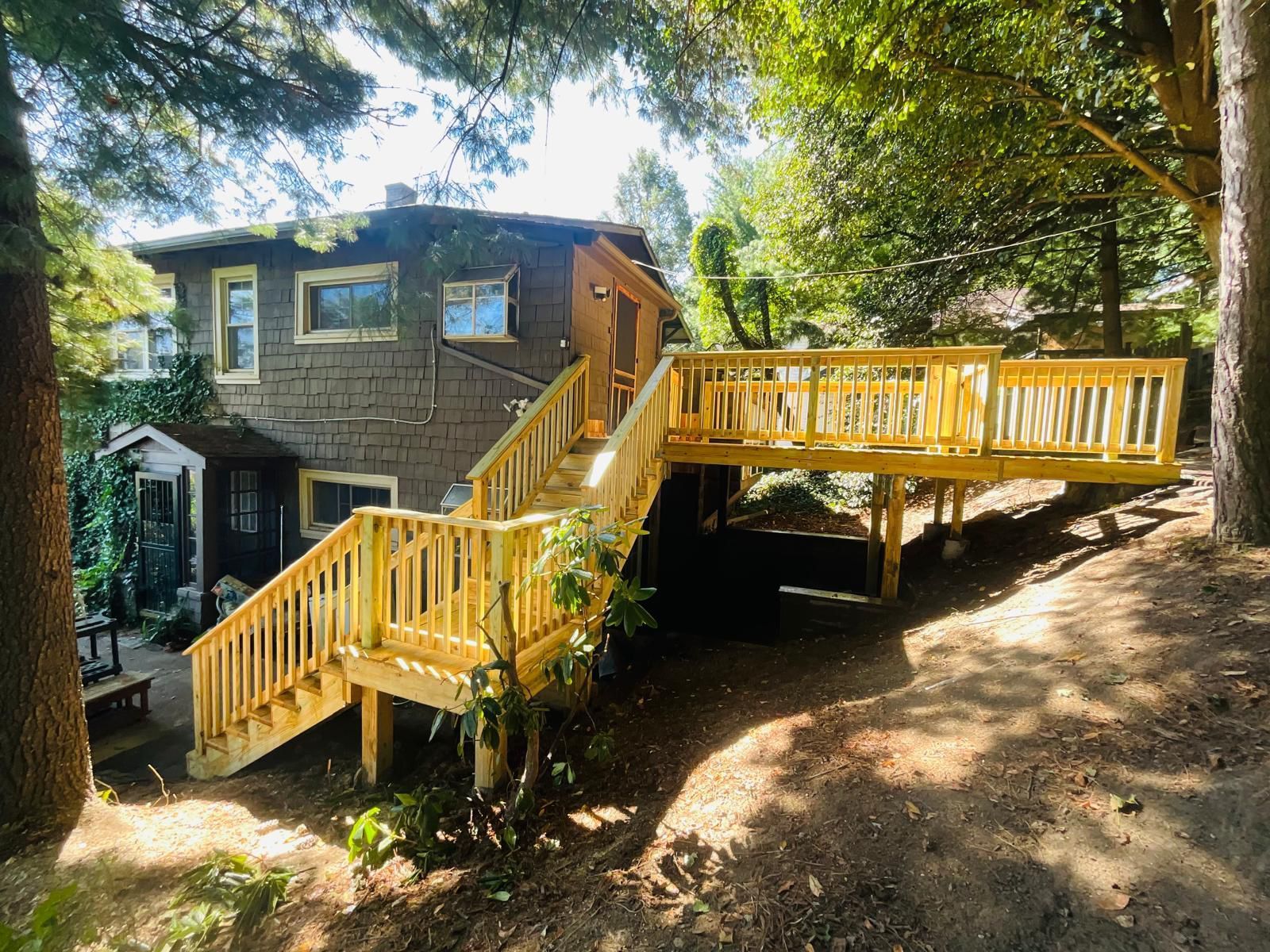 Wooden deck with stairs attached to a two-story house, surrounded by trees.