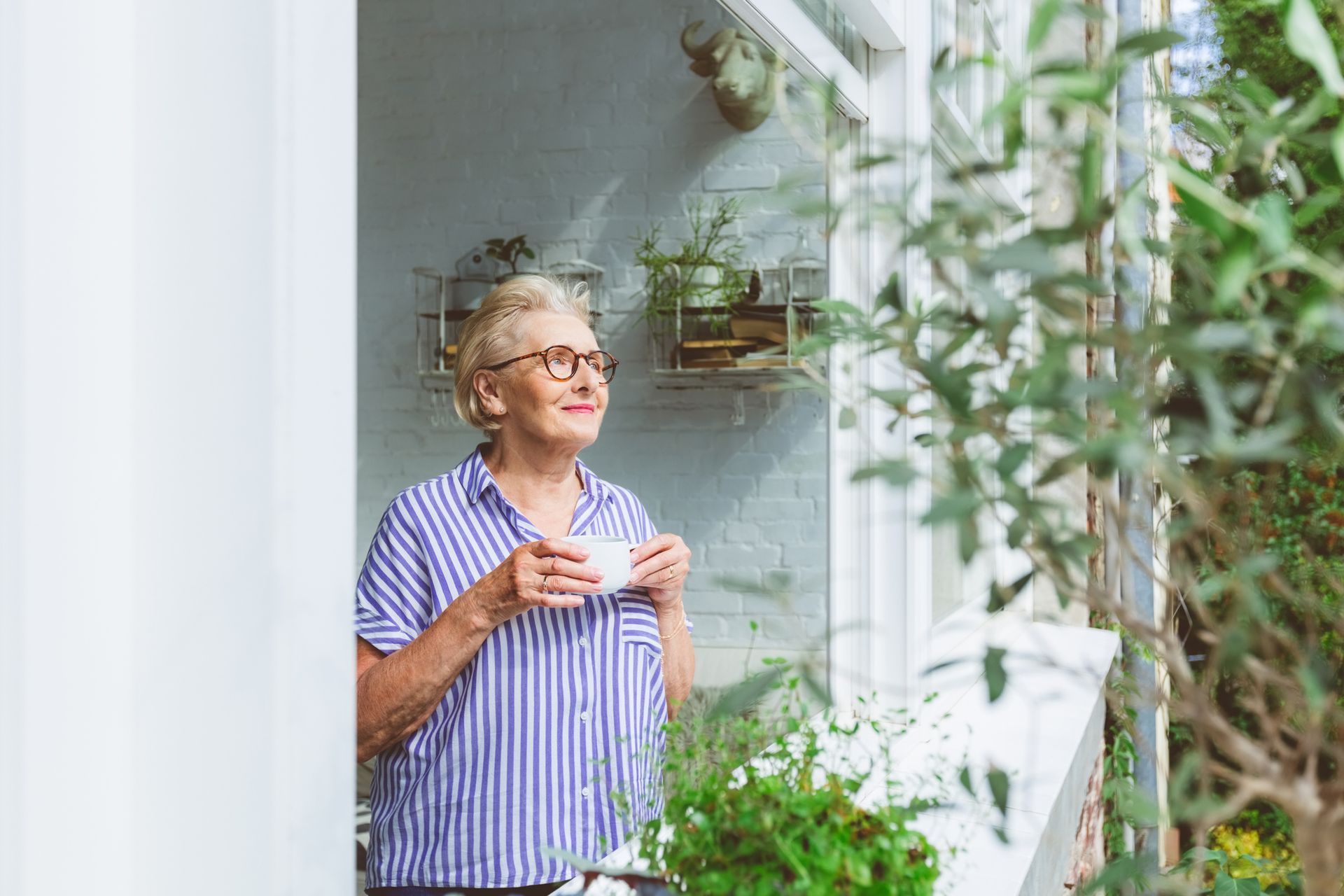 An elderly woman is looking through the window while having coffee at her senior housing apartment.