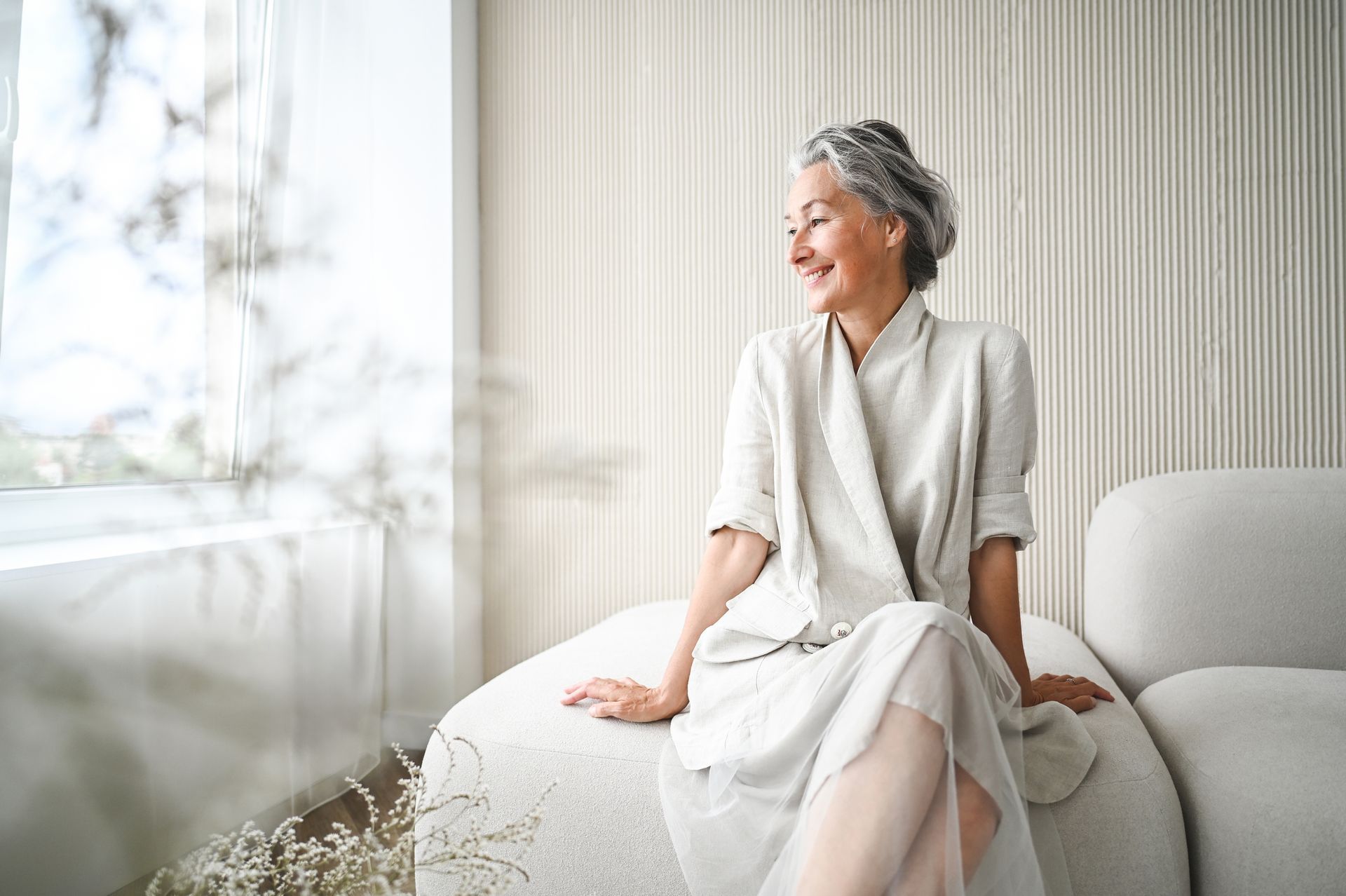 Woman with gray hair smiles while sitting on a white sofa near a window.