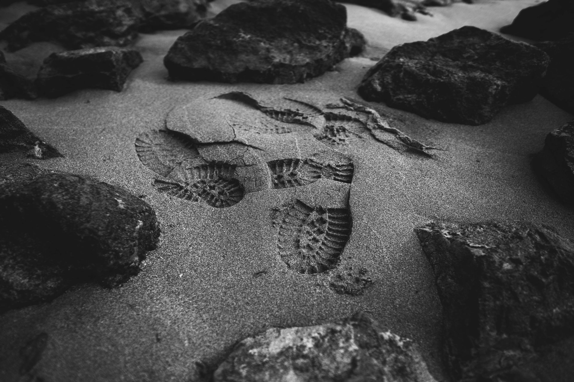Footprints in sand surrounded by rocks, in grayscale. SPATIAL FINE ARTS