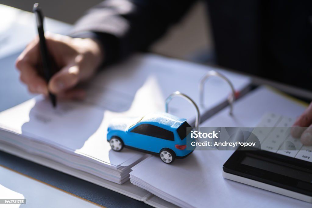 Person writing on paper, blue toy car on documents, calculator on desk.