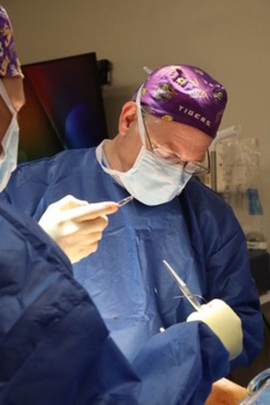 Surgeon in blue scrubs and a purple cap operating, holding tools, focused. Operating room setting.