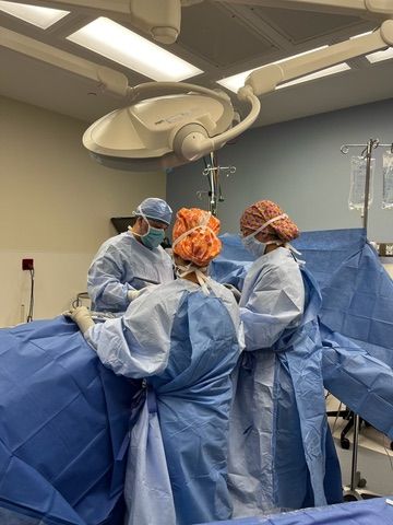 Surgeons in blue scrubs operate on a patient, illuminated by overhead surgical lights in an operating room.