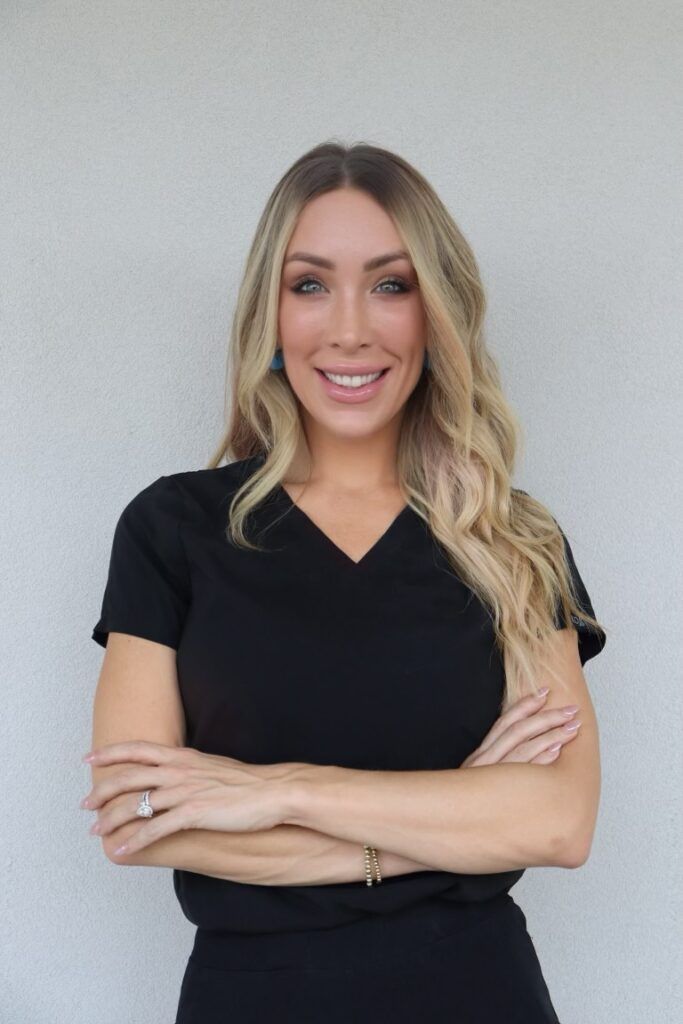 Blonde woman in black top smiles with arms crossed, standing in front of a neutral wall.