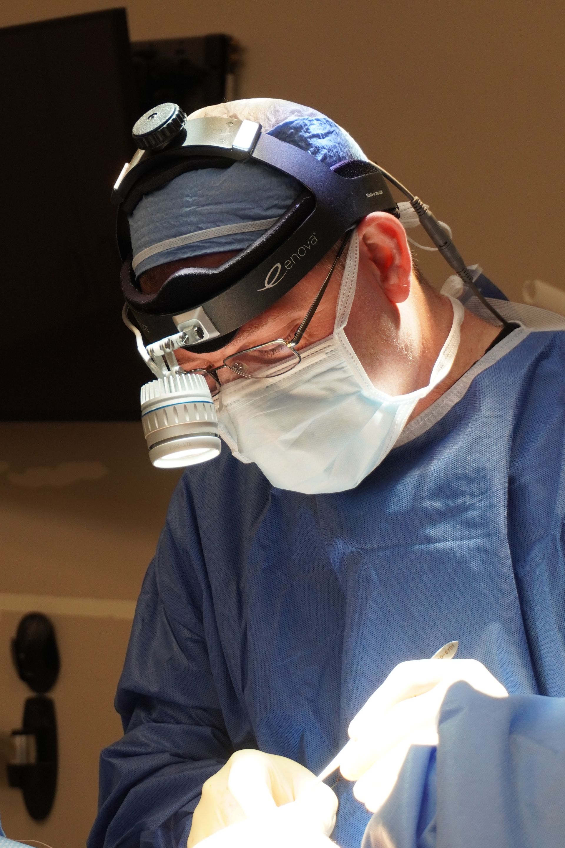 Surgeon in blue scrubs and mask, wearing a headlamp and glasses, operating.