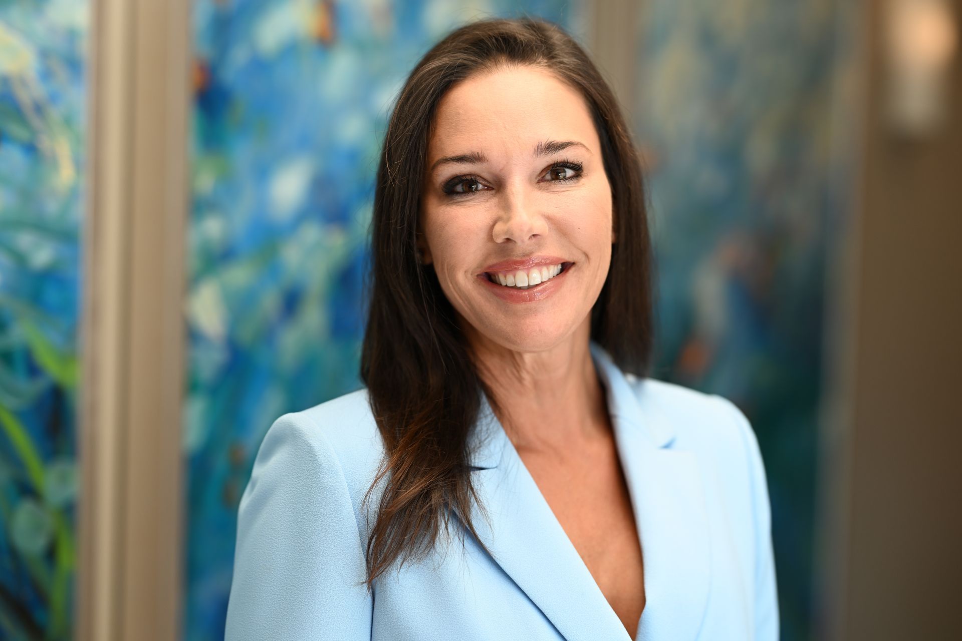 Woman in light blue blazer smiles in front of blue artwork.