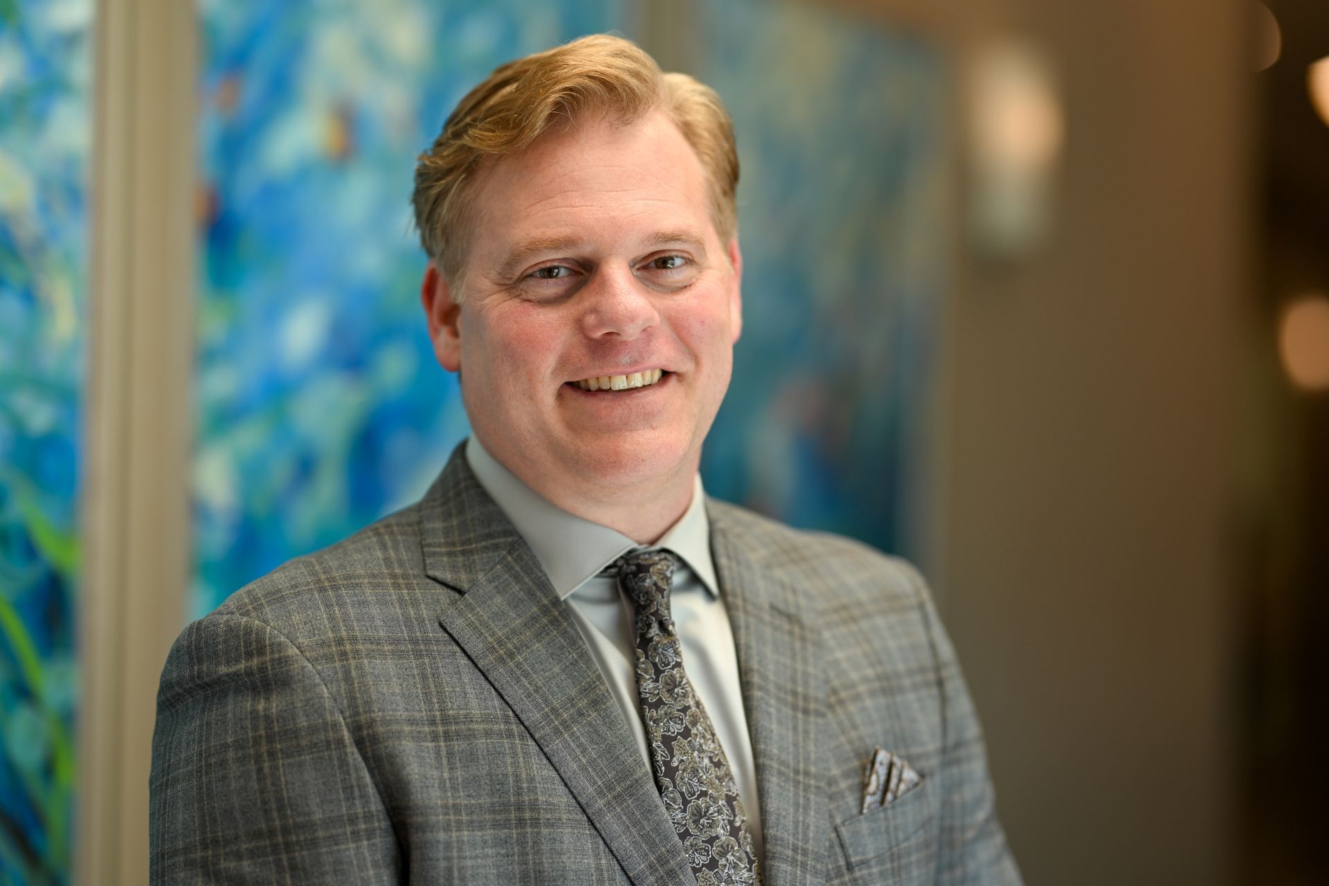 Man in gray plaid suit, smiling, in front of blue artwork.