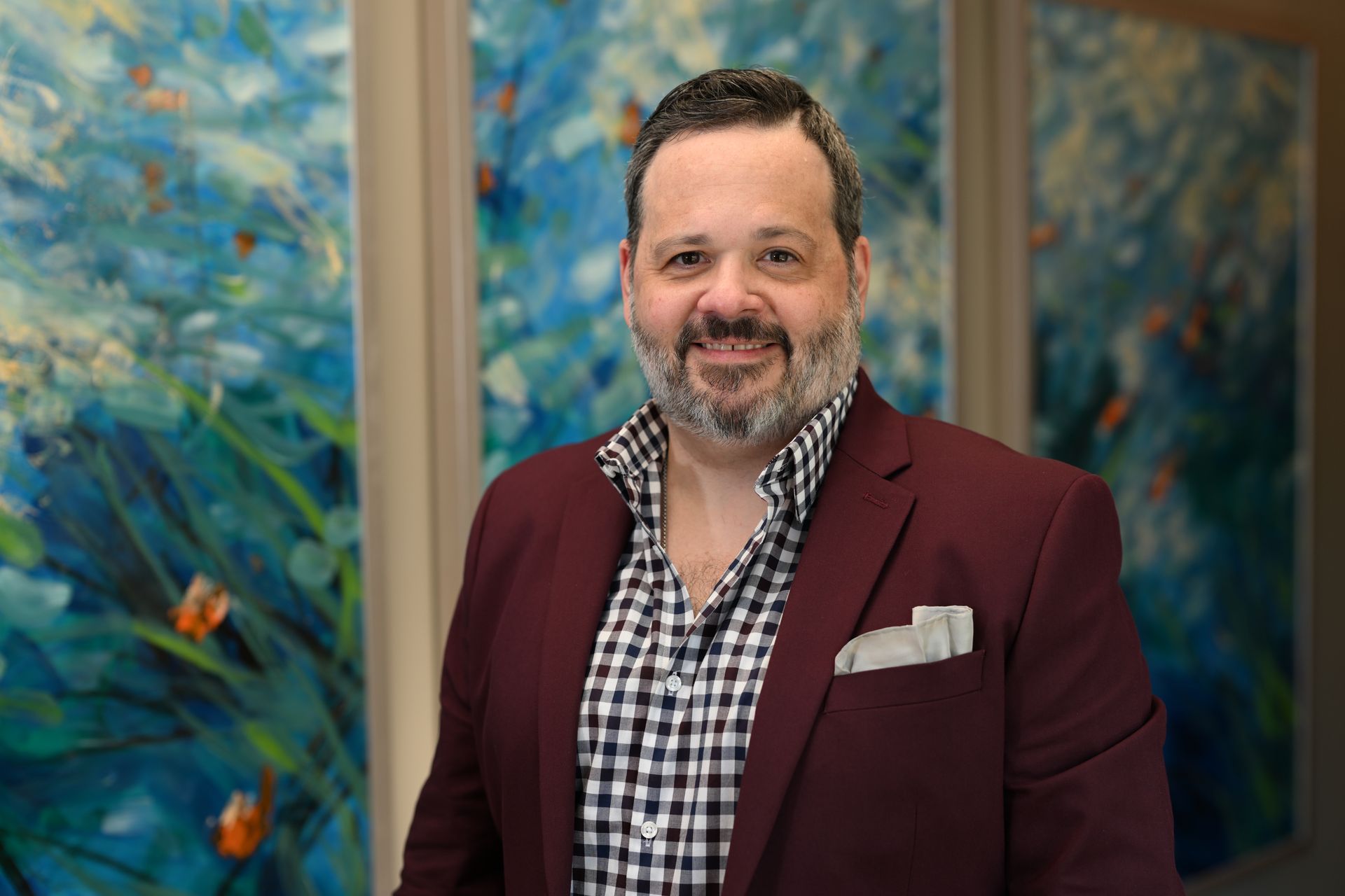 Man in burgundy blazer smiles, checkered shirt, standing in front of abstract blue and orange art.