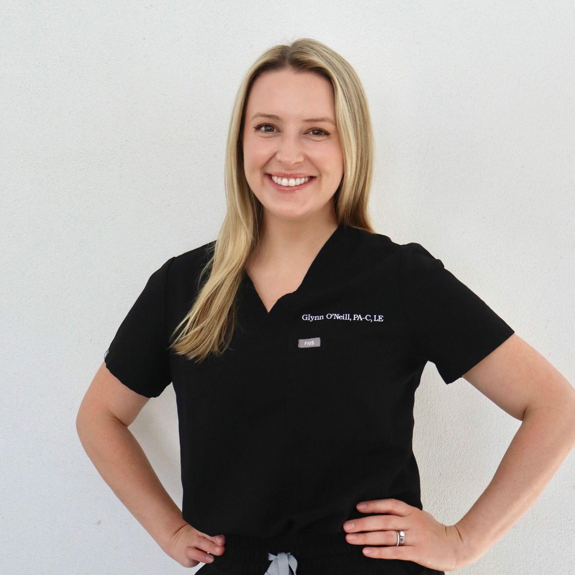 Woman in black scrubs, smiling, hands on hips, standing in front of a white wall.