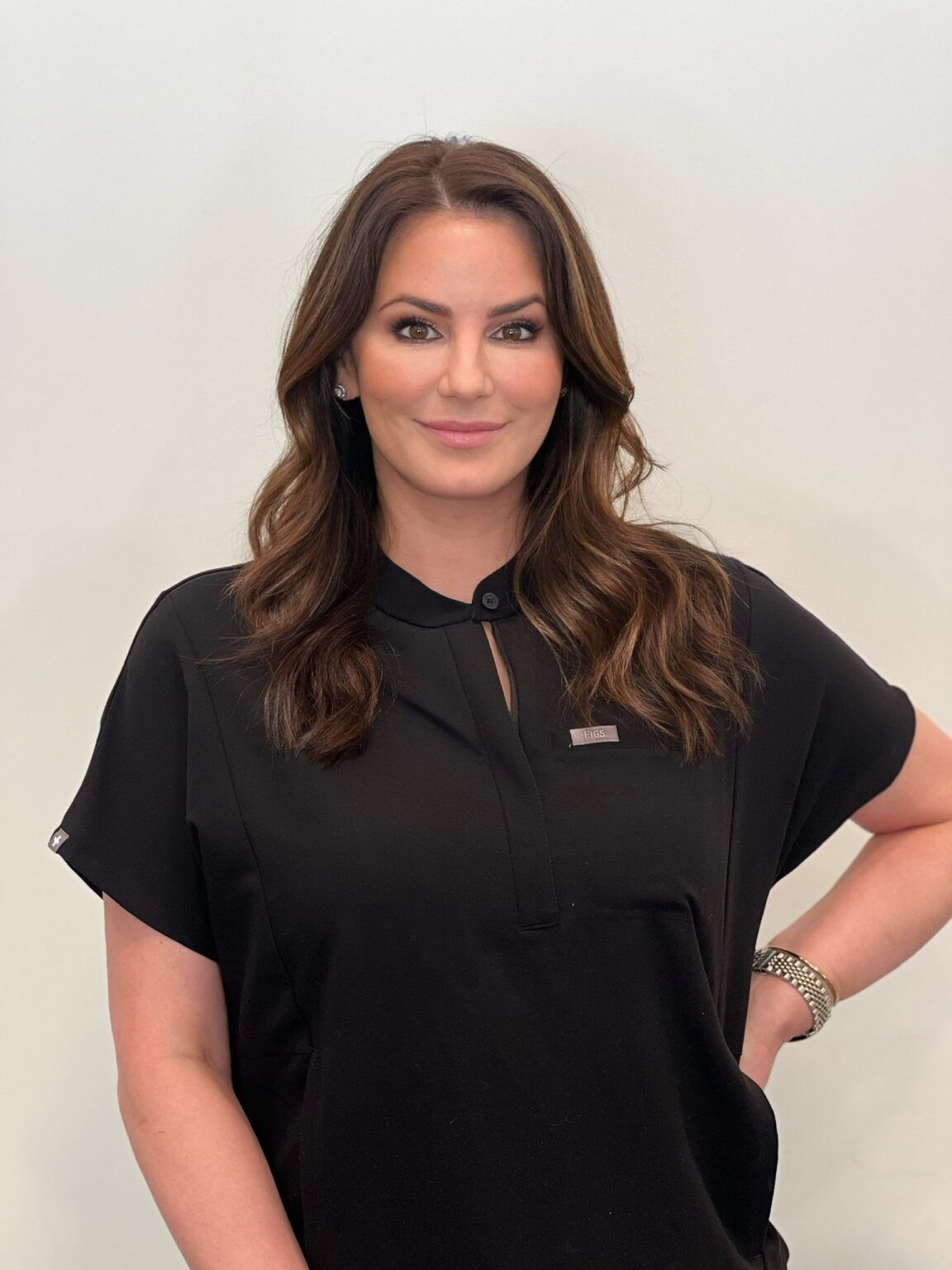Woman with long brown hair, wearing a black top, smiling, hand on hip, against a white wall.