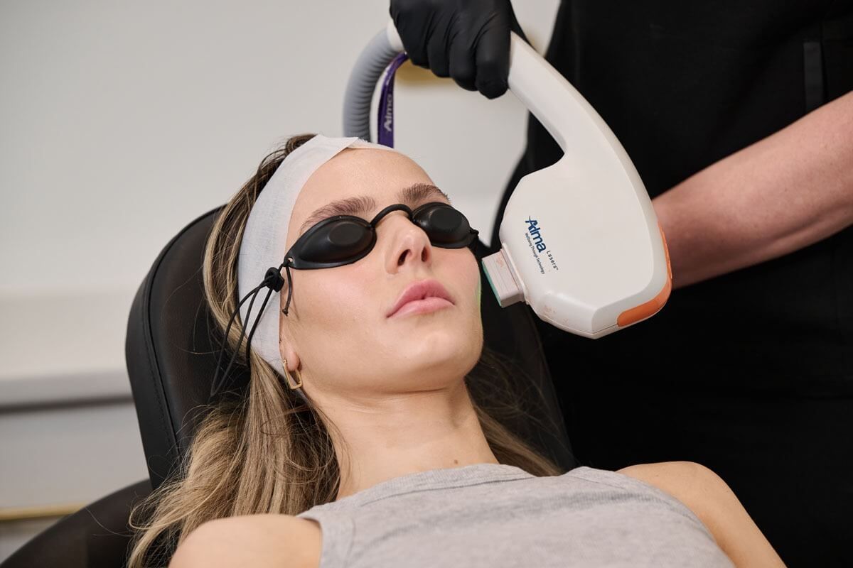 Woman undergoing laser facial treatment, wearing protective eyewear and head wrap in a clinic setting.