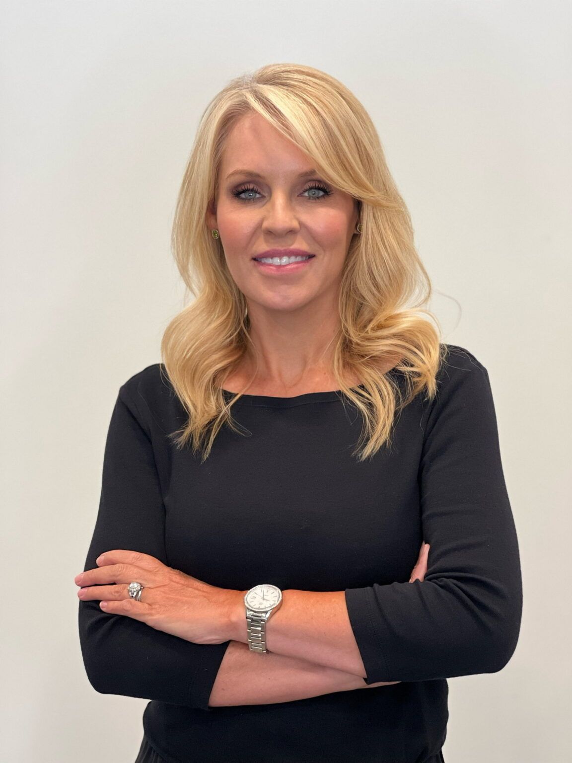 Woman with blonde hair, arms crossed, wearing a black top and a watch; smiling in front of a white background.