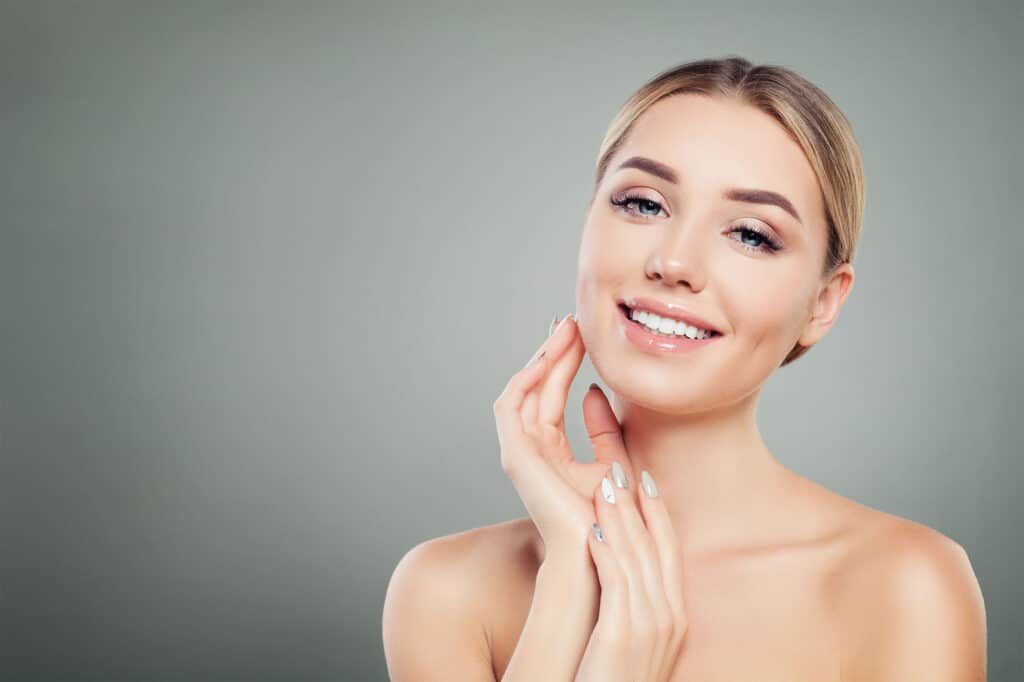 Woman with fair skin smiles, touching her neck and face, against a gray background.