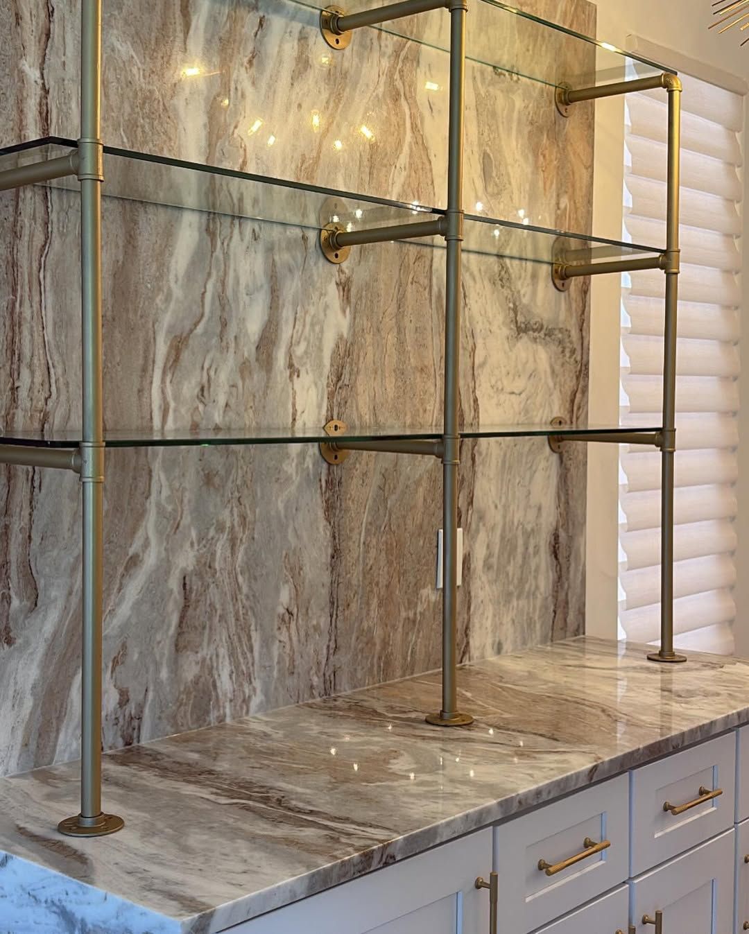 Marble-tiled shower with brass fixtures and built-in shelves above a white vanity with gold handles