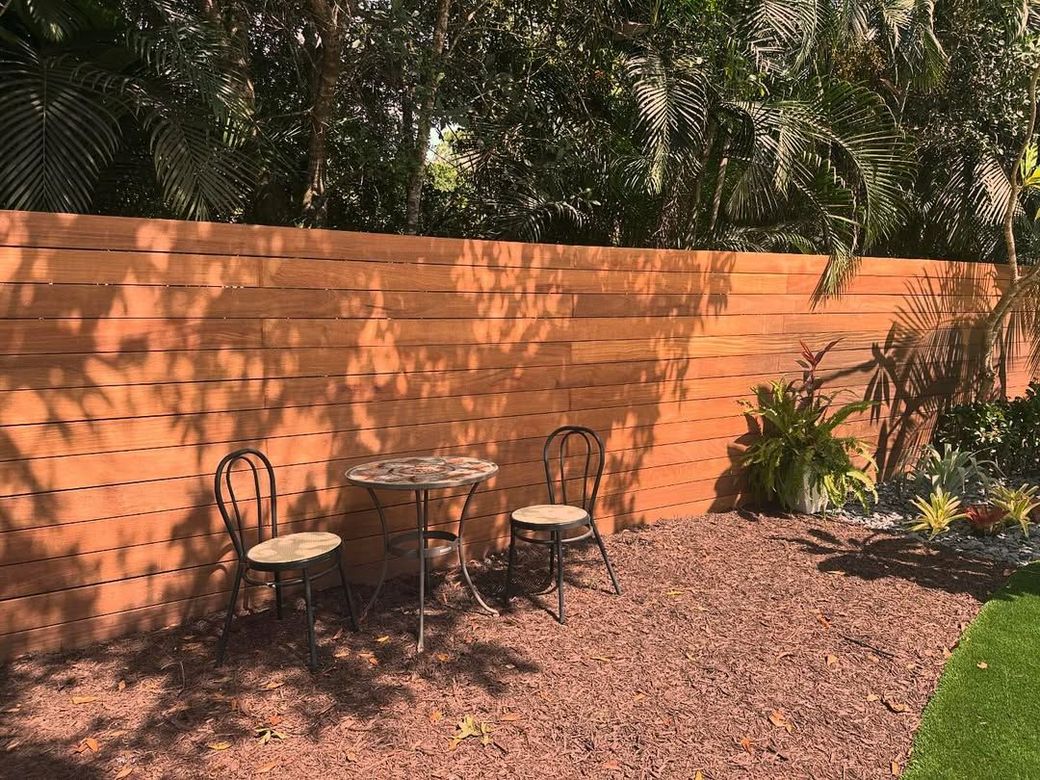 Small patio with two chairs and a table against an orange wall, surrounded by plants and gravel.