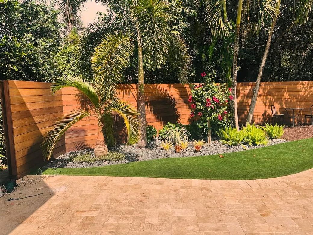 Backyard garden with curved lawn, wooden fence, palm trees, and flowering shrubs beside a patio