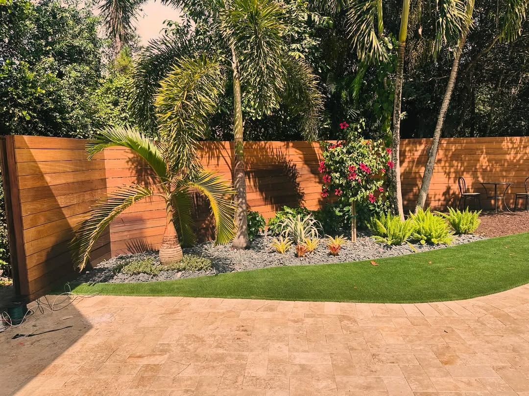 Backyard garden with curved lawn, wooden fence, palm trees, and flowering shrubs beside a patio