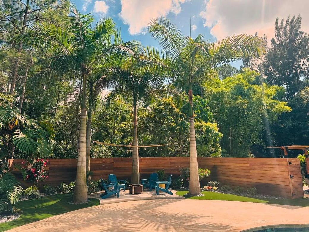 Sunny tropical patio with palm trees, lounge chairs, and a wooden fence under a blue sky