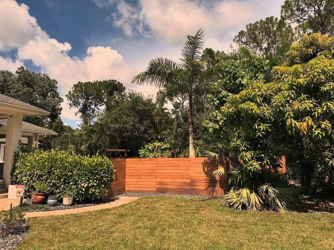 Sunny backyard with green lawn, orange fence, lush trees, and a partly cloudy sky