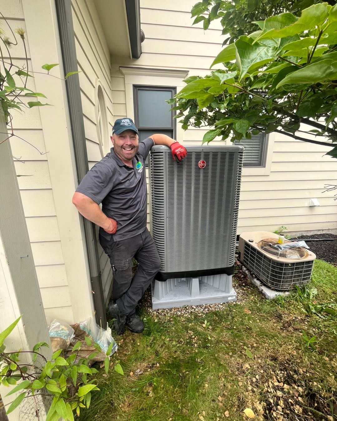HVAC technician stands by AC unit next to a house, smiling. Outdoors.