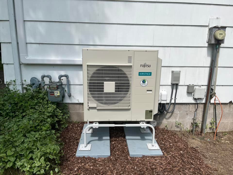 Fujitsu heat pump unit mounted on concrete pads beside a house wall and gas meter.