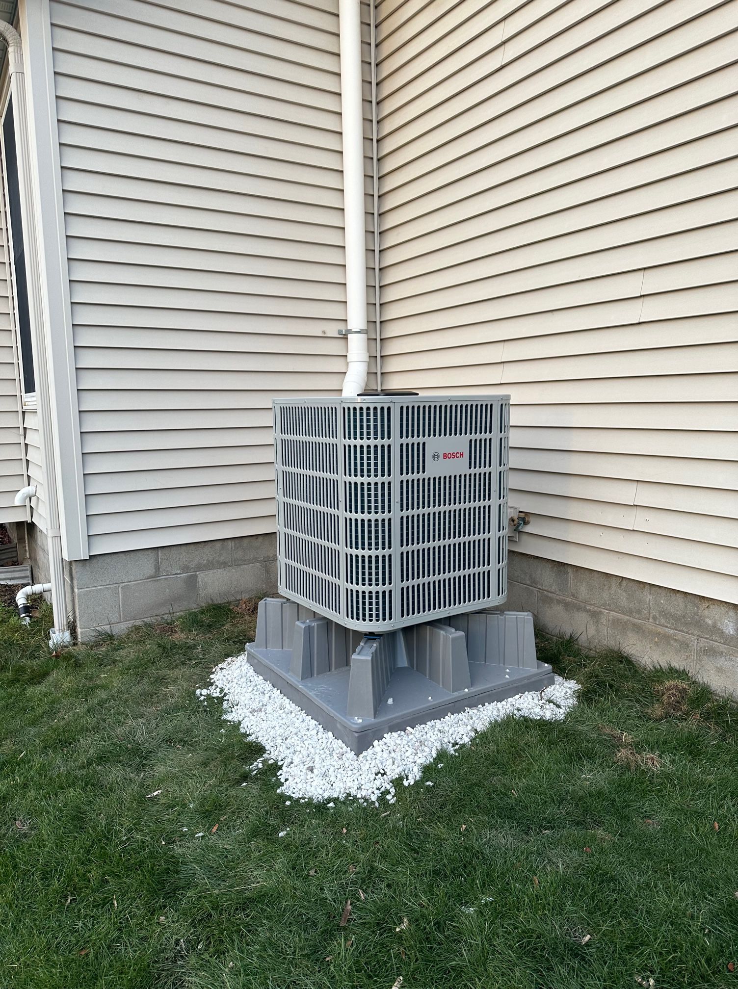 Air conditioner unit on a gray plastic base, beside a light-colored siding wall, with white gravel and green grass.